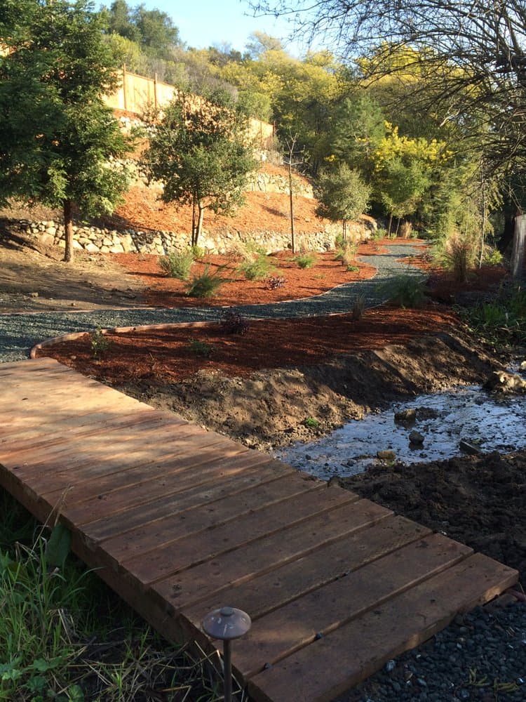 A wood bridge crossing a gravel dry-creek drainage swale on a landscaped Bay Area hillside