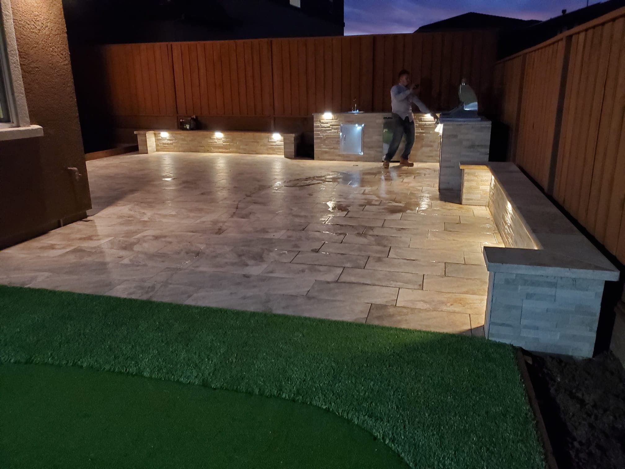 Travertine paver patio at dusk with illuminated stone seat walls, an outdoor kitchen, and artificial turf