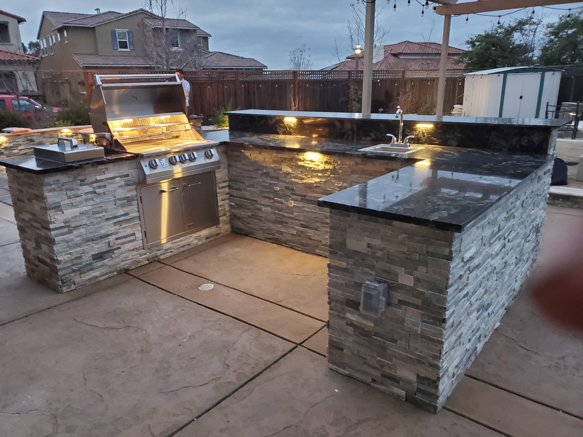 Stacked-stone outdoor kitchen island with a stainless grill and granite countertops lit at dusk